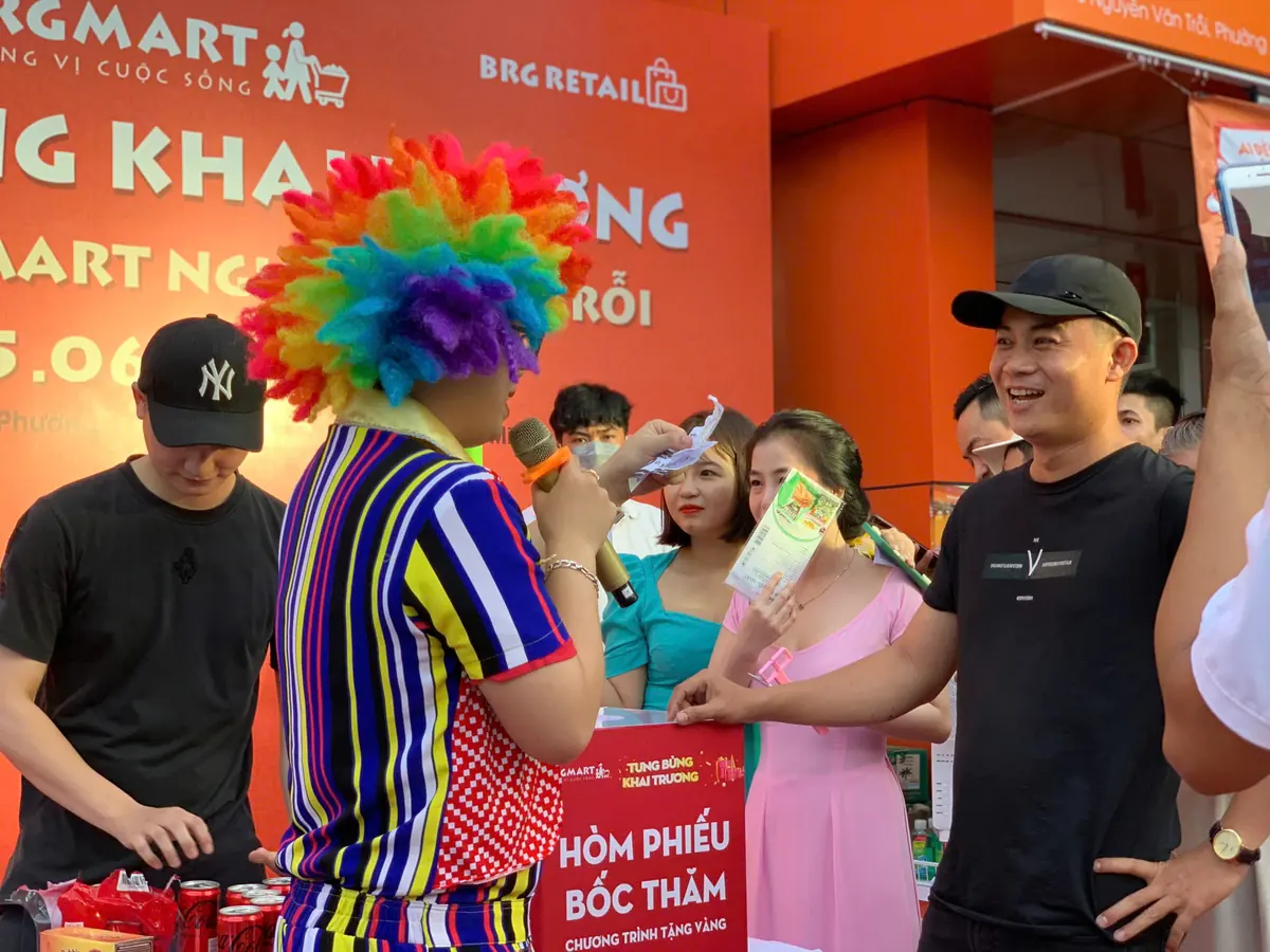 Hoạt náo viên hóa trang chú hề cầm micro, bốc thăm trúng thưởng cùng khách trước cửa siêu thị