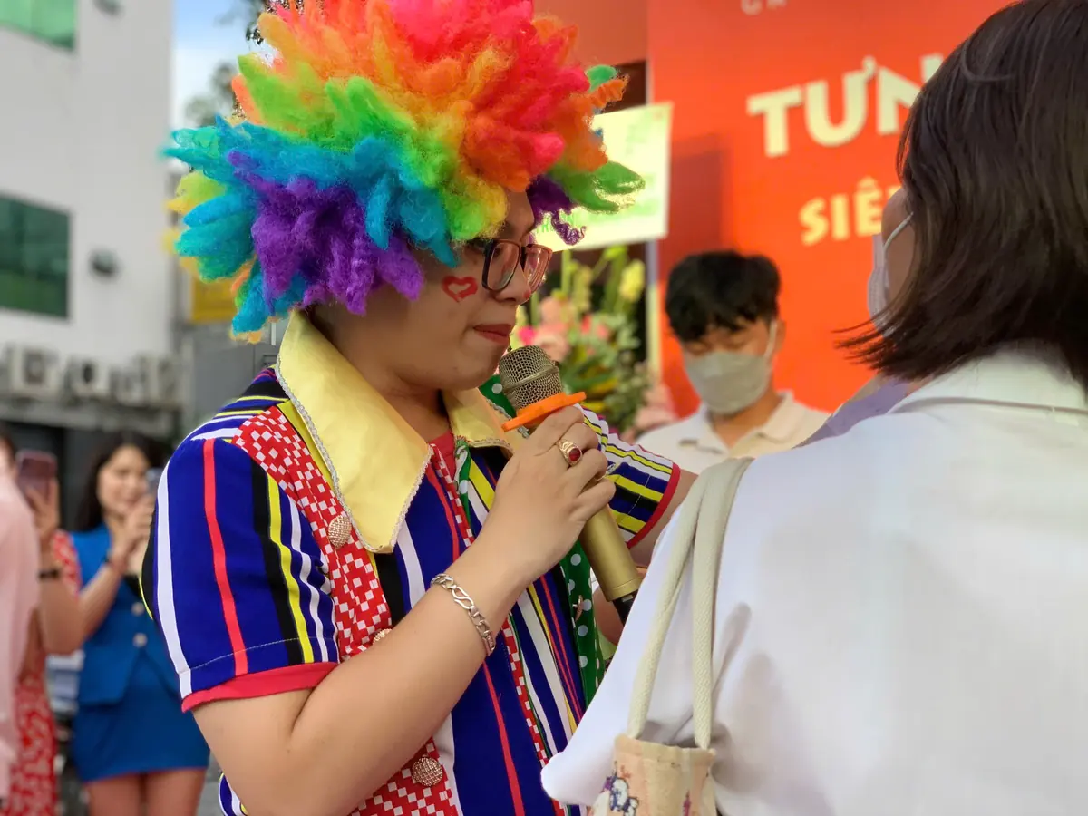Hoạt náo viên hóa trang chú hề đội tóc giả nhiều màu, cầm micro trò chuyện với khách ngày khai trương
