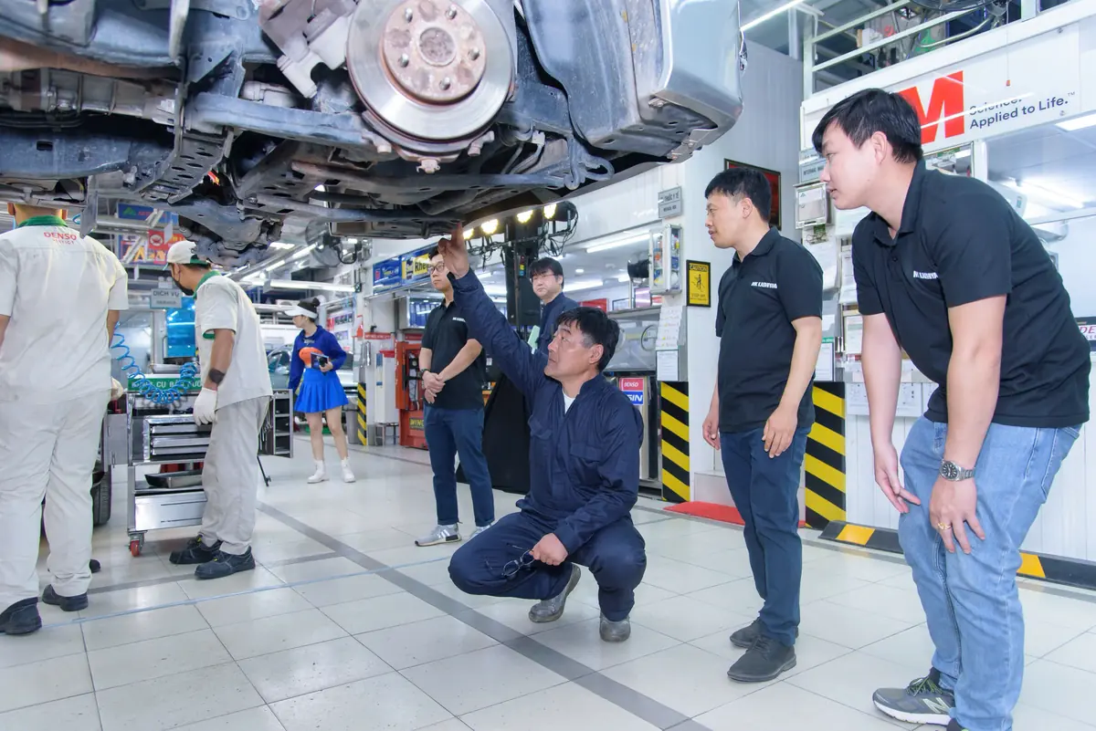 Chuyên gia ngồi xổm dưới gầm ô tô đang được nâng trên cầu, chỉ tay giải thích cho nhóm khách đứng quan sát trong xưởng dịch vụ